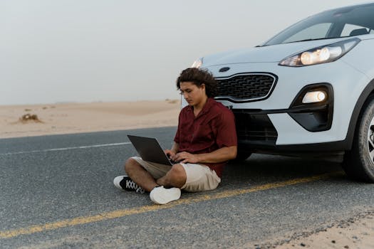 A man using a laptop while sitting on the road beside a parked car in a desert setting.