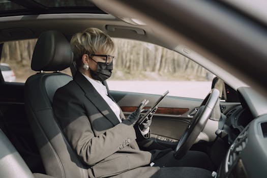 Side view of concentrated adult woman in formal clothes and protective mask and gloves sitting on driver seat of contemporary car and using tablet